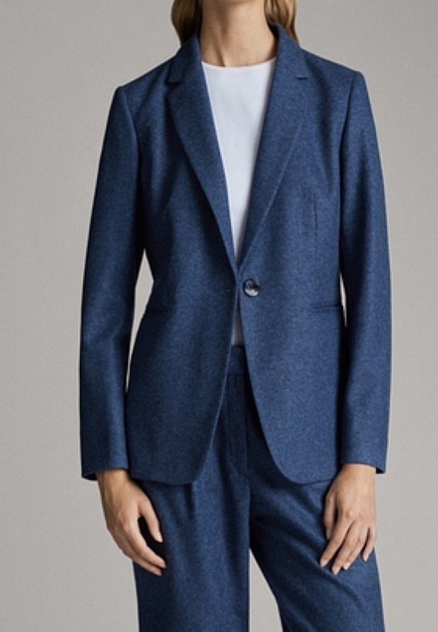 Femme portant un blazer bleu taillé sur mesure avec un bouton fermé et un pantalon assorti sur un haut blanc, debout devant un fond uni.