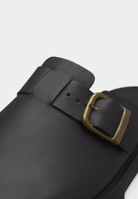 Μαύρο δερμάτινο σανδάλι με φαρδιά λωρίδα και ρυθμιζόμενο χάλκινο αγκράφα σε λευκό φόντο.