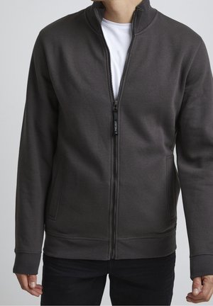 Homme portant une veste zippée gris foncé sur une chemise blanche, debout les bras détendus le long du corps devant un fond uni.