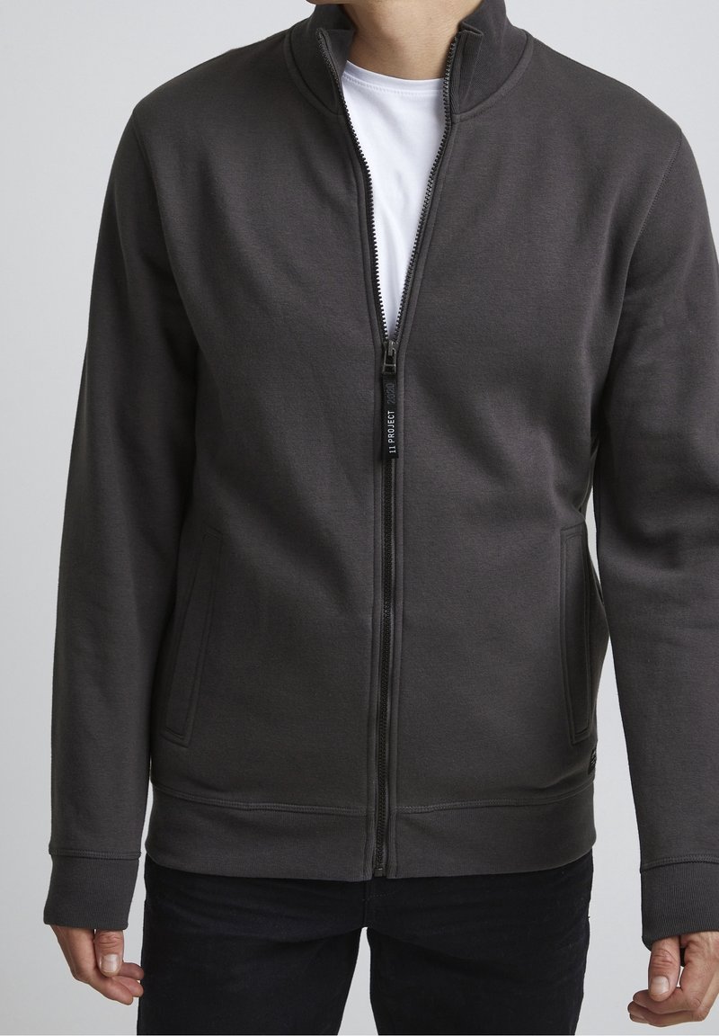Homme portant une veste zippée gris foncé sur une chemise blanche, debout les bras détendus le long du corps devant un fond uni.