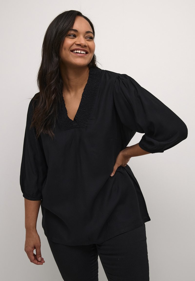 Schwarze Bluse mit V-Ausschnitt und gerüschtem Detail am Halsauschnitt, hat kurze, puffige Ärmel und eine lockere Passform. Dazu schwarze Hosen.
