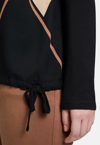 Schwarzes Langarmoberteil mit einem Kordelzugsaum, akzentuiert mit einem beigen und braunen geometrischen Design aus weichem Stoff.