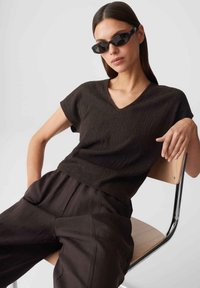 Woman wearing black sunglasses, a textured black short-sleeve top, and loose black pants, reclining on a wooden chair with metal legs.