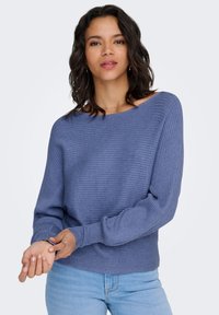 Maglione blu a coste con scollo ampio e maniche lunghe, realizzato in materiale morbido; abbinato a jeans azzurri.