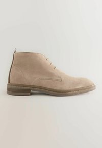 Beige Wildleder-Stiefeletten mit glatter Textur, ausgestattet mit einem niedrigen Absatz, minimaler Schnürung und einem kontrastierenden braunen Rückenpaneel.