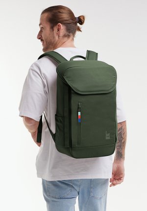 Muž se sepnutými vlasy, který má na sobě bílé tričko a džíny, nesoucí velký tmavě zelený batoh s bočními kapsami a předním zipem.