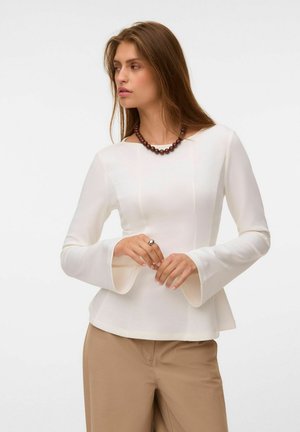 Femme aux longs cheveux bruns portant un haut blanc à manches longues avec des poignets évasés, un pantalon beige, un collier de perles foncé et une bague, regardant vers sa gauche.