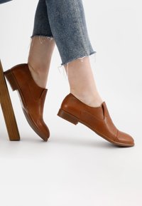 Bruine leren instapschoenen met een gladde afwerking, ronde neus, kleine hak en minimalistisch ontwerp, gecombineerd met versleten spijkerbroek.