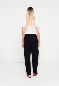 Femme aux longs cheveux blonds portant un haut blanc à manches courtes, un pantalon noir taille haute et des sandales rouges, debout face à un fond blanc.