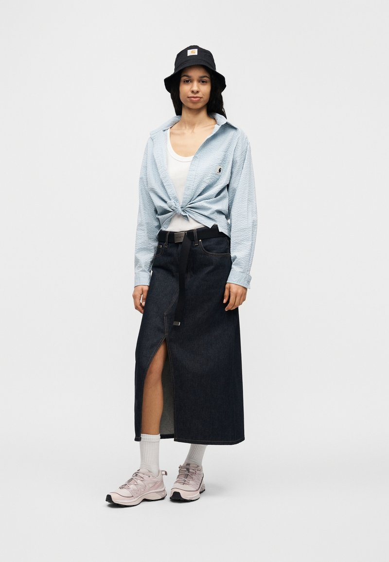 Chemise bleu clair à col, nouée à la taille, associée à une jupe longue en jean foncé avec une fente à l'avant. Portée avec des baskets roses et des chaussettes blanches.