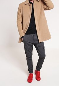 Beige fleecejacka med rund krage, svart skjorta, grå cargobyxor med elastiska muddar och starkt röda sneakers.