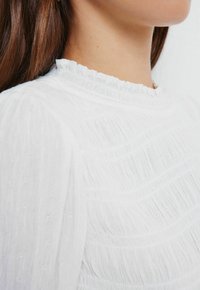 Bílá textura košile s volánkovým výstřihem, která obsahuje horizontální pruhy a jemnou, lehkou látku. Detailní pohled na rameno.