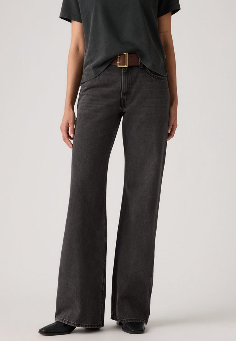 Άτομο που φοράει μαύρο τζιν με φαρδιά πόδια, μαύρη ζώνη με χάλκινη αγκράφα, μαύρο κοντομάνικο μπλουζάκι και μαύρα παπούτσια, στέκεται μπροστά σε απλό φόντο.