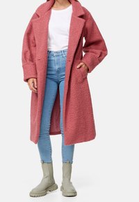 Manteau rose à texture duveteuse avec un col à revers, deux poches latérales, et une coupe mi-longue, porté par-dessus un jean slim bleu clair et des bottes grises.