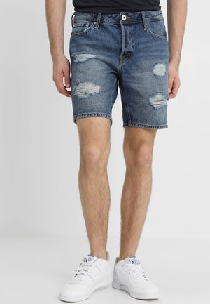 Man som bär blå slitna denimshorts och vita sneakers, stående mot en ljus enfärgad bakgrund.