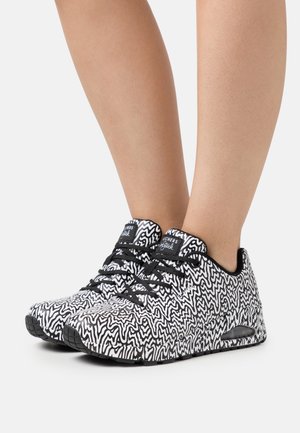 Gambe che indossano sneakers a motivi bianco e nero con lacci neri e suola imbottita su sfondo grigio chiaro.