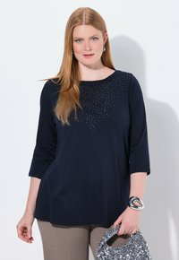 Top blu navy a tre quarti con scollo rotondo, decorato con strass disposti in un motivo gradiente, abbinato a pantaloni grigi.