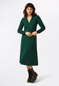 Grünes Kleid mit Punkten, langen Ärmeln und einem Kragen, mit einer gebundenen Taille. Getragen mit schwarzen Schnürstiefeln.