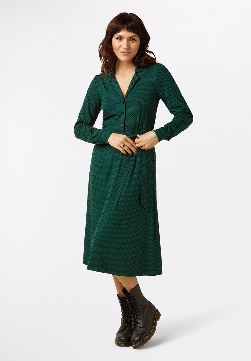 Grünes Kleid mit Punkten, langen Ärmeln und einem Kragen, mit einer gebundenen Taille. Getragen mit schwarzen Schnürstiefeln.