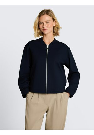 Blonde Frau trägt eine marineblaue Jacke mit Reißverschluss über einem gestreiften Oberteil und beigefarbenen plissierten Hosen, steht mit den Händen in den Taschen vor einem schlichten Hintergrund.
