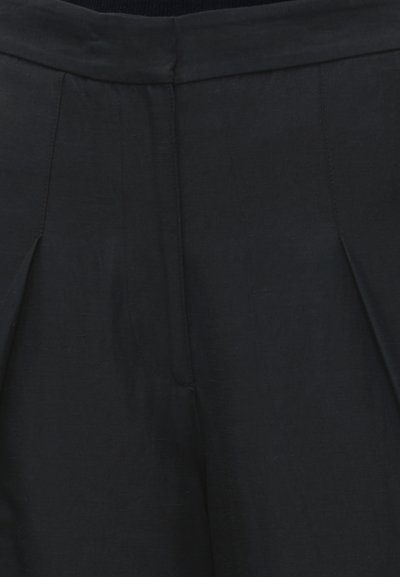 Pantalon noir taille haute avec plis sur le devant et une ceinture nette, montrant un gros plan du haut de la cuisse et de la zone de la taille.