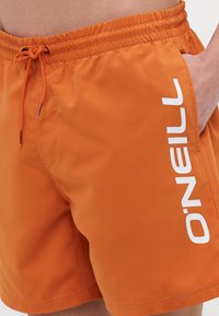 Oranssit uimapuvut, joissa on joustava vyötärö, kiristysnauha ja valkoinen graafinen logo sivulla. Sileä, kevyt rakenne.