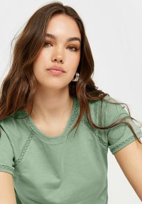 Maglietta verde chiaro con scollatura rotonda, maniche corte e dettagli in pizzo lungo le cuciture. Tessuto di qualità liscia e vestibilità casual.