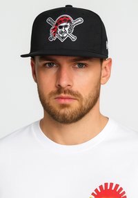 Schwarze Baseballmütze mit gesticktem Piratenmotiv, das ein rotes Bandana und gekreuzte Schläger zeigt. Getragen mit einem schlichten weißen Shirt.
