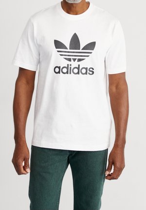 Mężczyzna w białej koszulce Adidas z czarnym logo trefoil i napisem, w ciemnozielonych spodniach, na jednolitym tle.