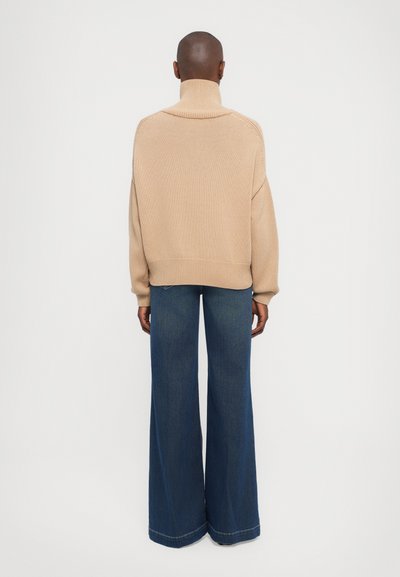 Beige Strickpullover mit hohem Kragen, lockerer Passform und geripptem Saum, kombiniert mit weiten, dunkelblauen Jeans.
