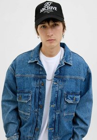 Denim jacket in light blue with a relaxed fit, featuring chest pockets and a faded texture, paired with a black cap displaying white logo.