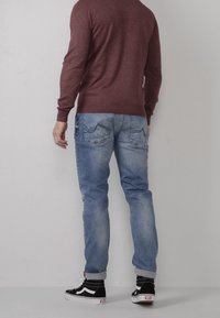 Persona vestita con un maglione bordeaux, jeans azzurri arrotolati e sneaker alte nere, in piedi contro un muro chiaro uniforme.