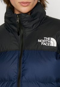 Polstret jakke fra The North Face, med en marineblå og svart fargepalett, høy krage, glidelås-lukking og logodetalj på brystet.