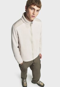 Helles beiges Fleece-Jackett mit hohem Kragen, Reißverschluss und Seitentaschen, kombiniert mit olivgrünen Cargo-Hosen und braunen Wanderschuhen.