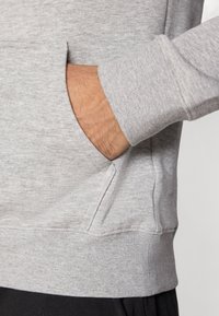 Hall halli pusa tekstuurse kangaga, millel on kängurutasku ja soonikkoes mansetid. Lähedalt vaadatuna tasku ja varruka detailid.