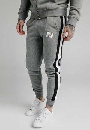 Man met grijze joggingbroek met zwart-witte strepen aan de zijkant, witte sneakers en getatoeëerde handen tegen een effen grijze achtergrond.