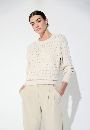 Mujer con un jersey crema con pequeñas tachuelas doradas y pantalones beige de talle alto, de pie con las manos en los bolsillos contra un fondo blanco.