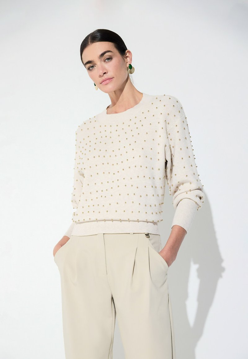 Femme portant un pull crème avec de petites clous dorés et un pantalon beige taille haute, debout les mains dans les poches devant un fond blanc.