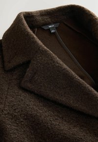 Manteau en laine marron avec une surface texturée, présentant un col pointu, une fermeture éclair dissimulée et une doublure intérieure douce. Étiquette de marque visible.