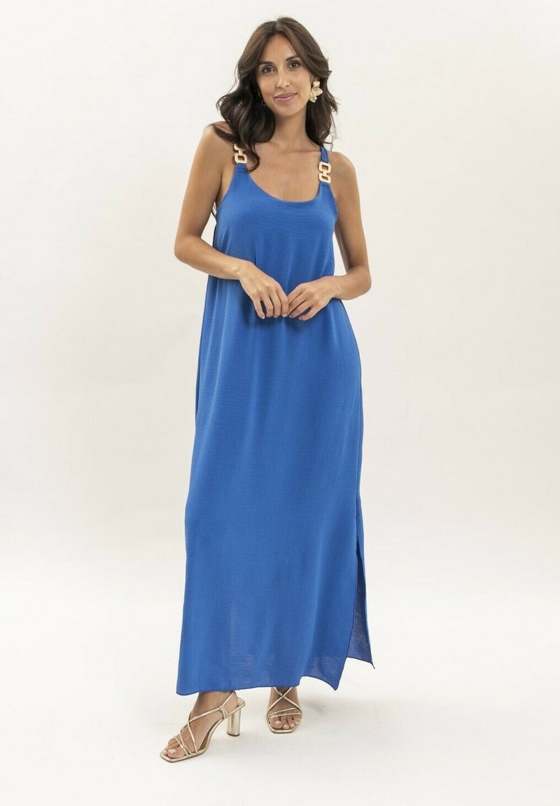 Robe maxi bleue avec larges bretelles présentant un grand matériel décoratif. Fabriquée en tissu lisse, elle comprend une fente sur le côté et tombe jusqu'aux chevilles.