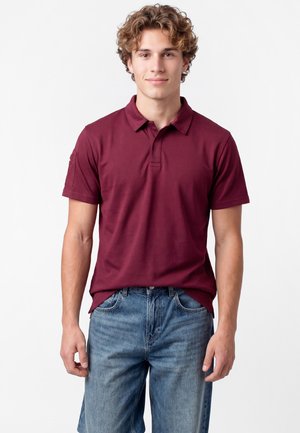 Junger Mann mit lockigem Haar, der ein bordeauxfarbenes Poloshirt und blaue Jeans trägt, steht vor einem schlichten weißen Hintergrund.