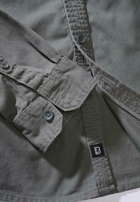 Szürke ing részlet varrott varrásokkal, kerek gombokkal és egy fekete alapon fehér "B" betűs címkével. Texturált szövet sima felülettel.