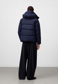 Osoba widziana od tyłu, ubrana w granatową kurtkę puchową z kapturem i luźne czarne spodnie o szerokich nogawkach na jednolitym jasnym tle.