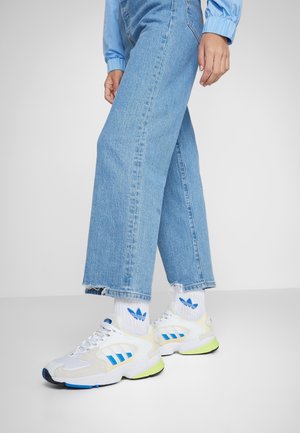Person iført lyseblå vide jeans, hvide Adidas-sokker og hvide sneakers med blå og neon grønne detaljer, stående på en grå gulv.