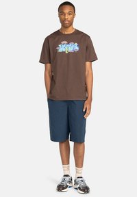 Bruine katoenen t-shirt met grafische print op de voorkant, gecombineerd met marineblauwe shorts. Model draagt witte sneakers met zwarte en rode accenten.