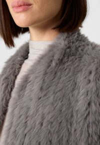 Grau gefärbtes Kunstfellkleidungsstück mit einer weichen, strukturierten Oberfläche und einem breiten Kragen. Das darunterliegende Outfit besteht aus einem leichten, glatten Stoff in neutralem Farbton.