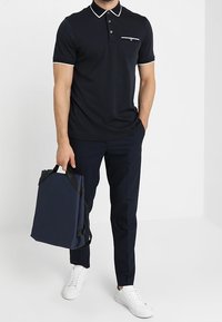 Granatowa koszula polo z białym wykończeniem kołnierzyka i kieszenią na piersi, połączona z granatowymi spodniami i białymi trampkami. Trzyma ciemnoniebieską torbę na ramię.