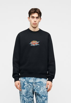 Ung mand iført sort sweatshirt med flaming Santa Cruz-logo og blå mønstrede jeans, stående mod en ensfarvet lys baggrund.