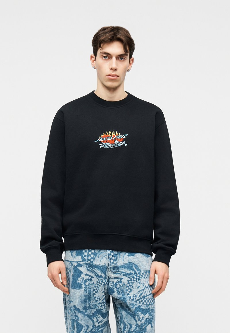 Νεαρός άνδρας που φοράει μαύρο φούτερ με το λογότυπο Santa Cruz σε φλόγες και μπλε τζιν με σχέδια, στέκεται μπροστά σε απλό ανοιχτό φόντο.