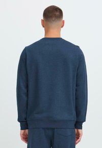 Rövid hajú férfi, aki sötétkék pulóvert és azonos színű nadrágot visel, háttal áll egy egyszerű fehér háttér előtt.